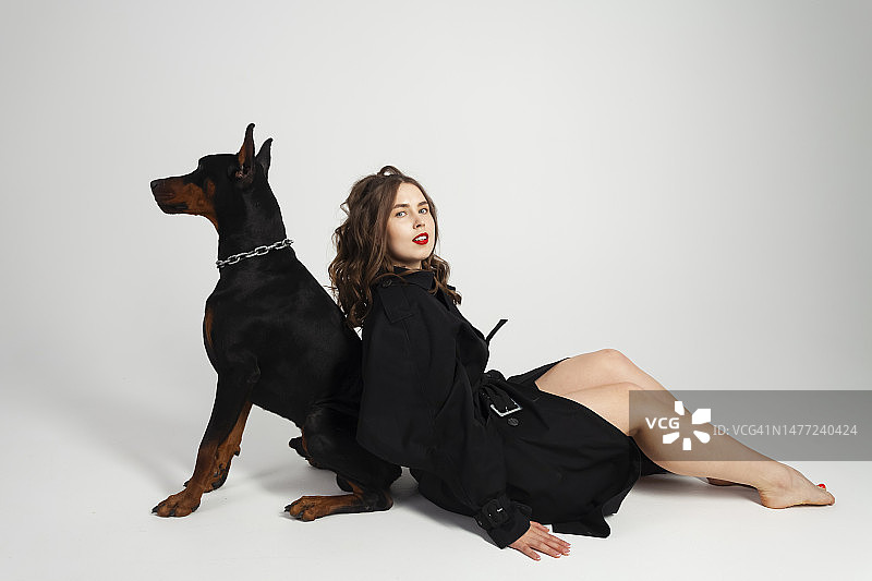 年轻漂亮的杜宾犬，脖子上戴着链子，主人是一位穿着黑色长外套、涂着鲜红色口红的优雅女士，白色背景隔离图片素材