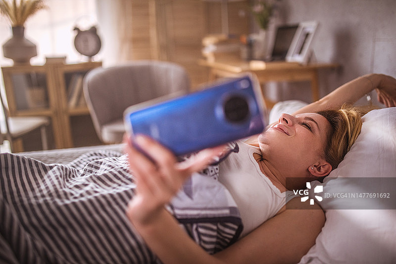 在床上醒来时使用智能手机的年轻女人图片素材