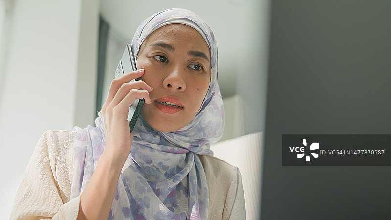 快乐的年轻穆斯林女性坐在床上，用电脑和手机与同事交谈工作特写。在家工作。图片素材