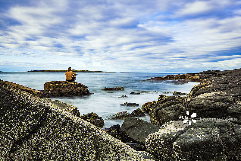 年轻的半裸男子坐在海边的岩石上，眺望着地平线图片素材