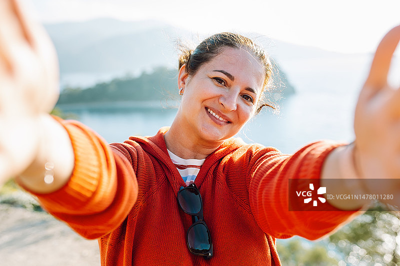 面带笑容的快乐女人在户外以大海和山脉为背景的自拍图片素材