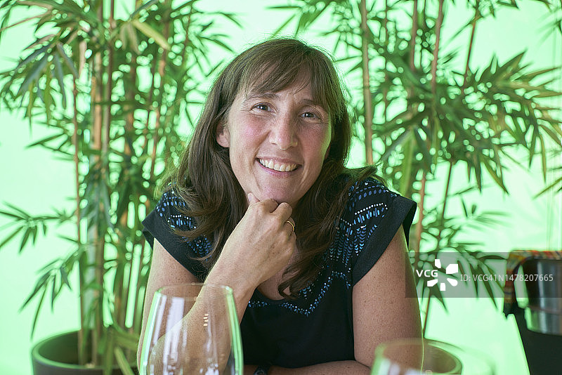 在餐厅里，面带微笑坐在桌旁，手持两杯葡萄酒，看向镜头的女子肖像图片素材