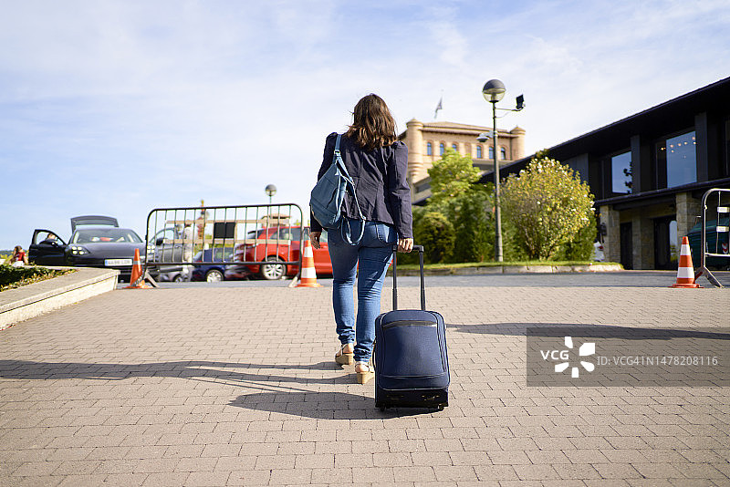 女子穿着夹克，背着背包，拉着行李箱走在街上的背影，背景是停放的汽车图片素材