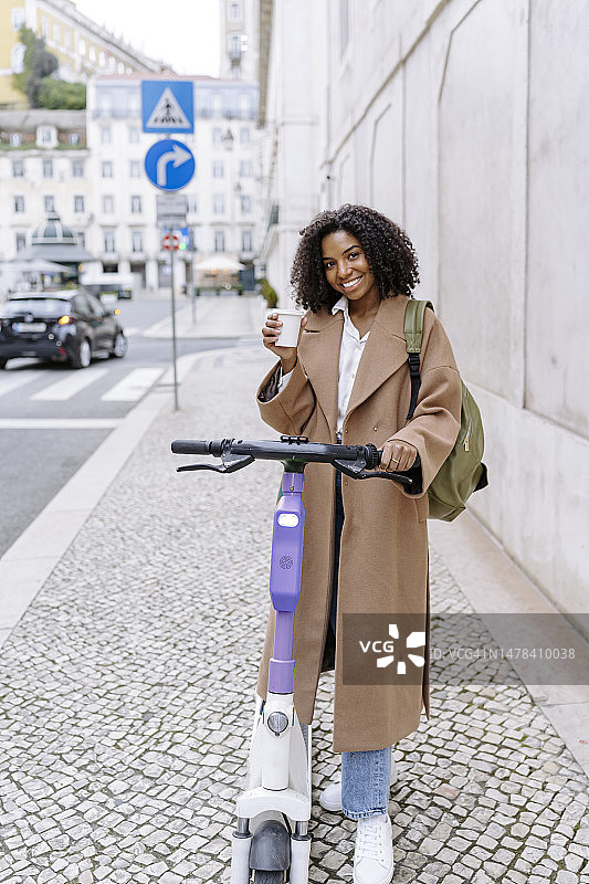 快乐的年轻女子手持咖啡杯和电动滑板车站在人行道上图片素材