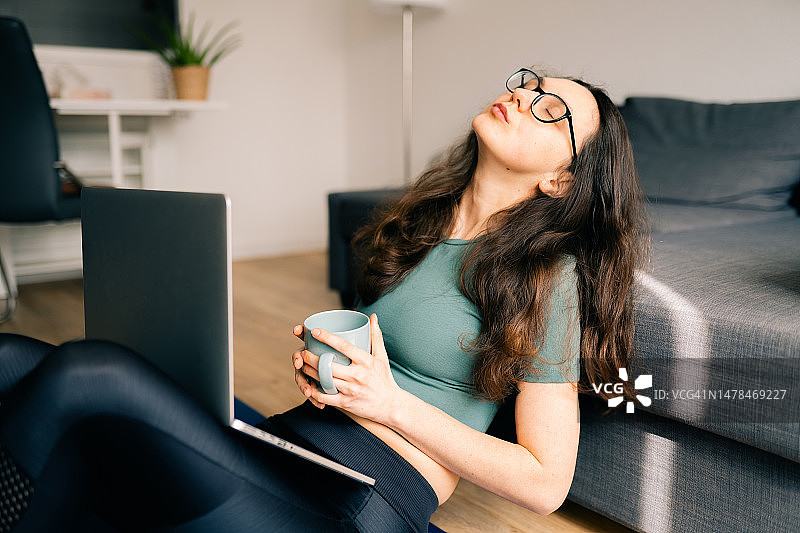 年轻的棕发女人在家工作时，拿着咖啡杯和笔记本电脑感到疲倦图片素材