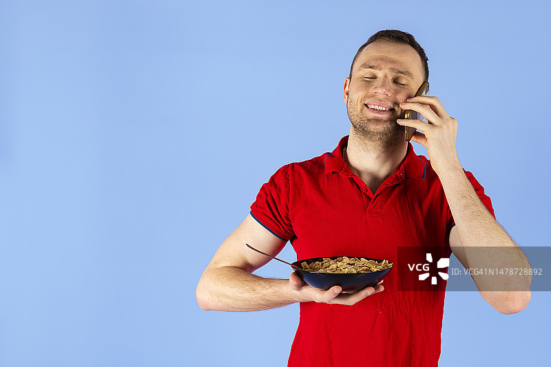 年轻英俊健康的男人穿着红色POLO衫，拿着一碗麦片或早餐，正在打电话。蓝色背景图片素材
