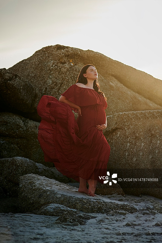 孕期海滩孕妇照：日落时分，准妈妈在海边岩石旁摆姿势，穿着时尚，手托腹部图片素材