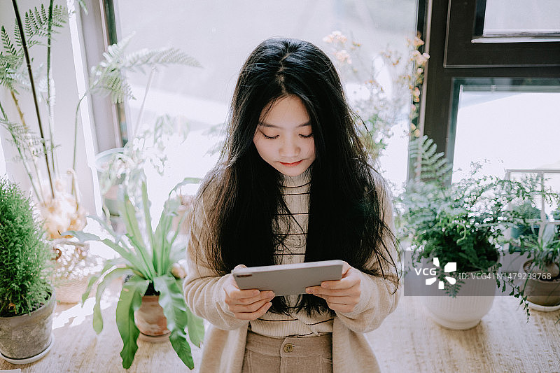 在摆满盆栽植物的窗台上使用平板电脑的年轻亚洲女性图片素材