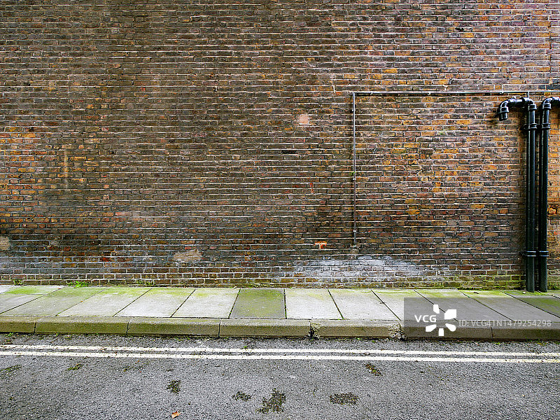带有黑色管道的风化棕色砖墙，混凝土人行道和带有标记的沥青街道，伦敦，英国图片素材