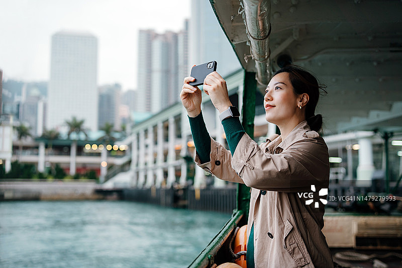 年轻亚洲女性在香港天星小轮上用手机拍摄城市天际线图片素材