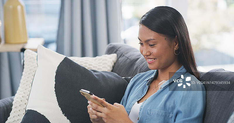 快乐的女人在沙发上使用智能手机，在休息室阅读社交媒体帖子、在线聊天和搜索。亚洲女性在房子里用手机打字，微笑和下载手机游戏图片素材