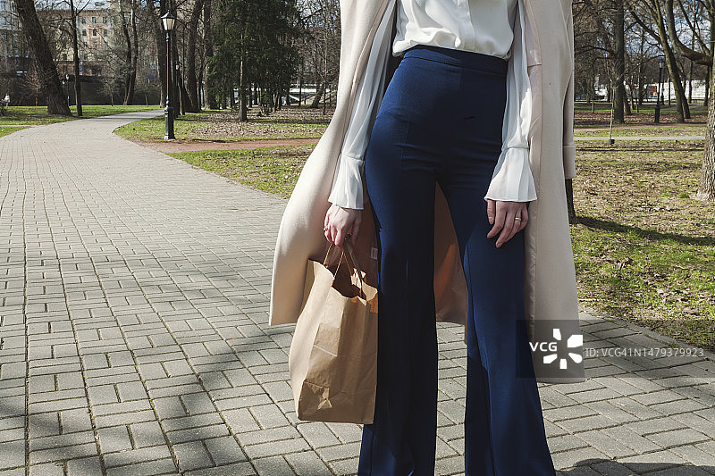 在公园里的时尚女孩，披着外套，手腕上戴着智能手表，穿着时髦的紧身深蓝色裤子和白色衬衫，拿着纸袋图片素材