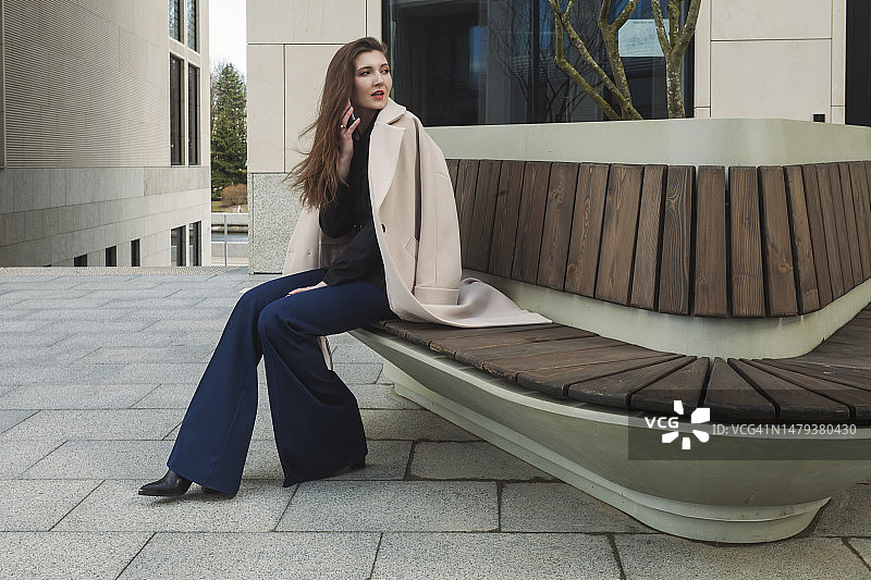 披着白色外套坐在商业区长椅上打电话的美丽女商人图片素材
