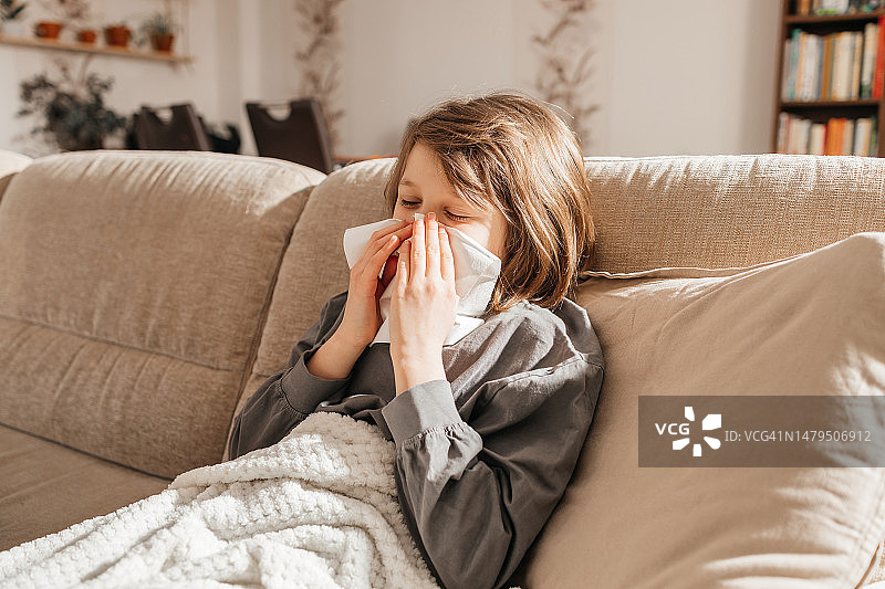 生病的少女坐在沙发上，把鼻子擤进纸手帕里。儿童得了流感图片素材