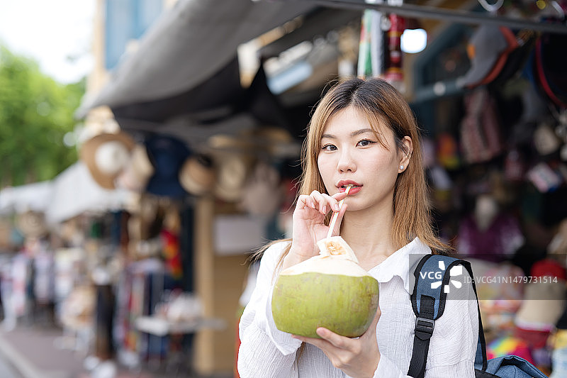 旅行时保持水分：快乐的韩国女孩在曼谷街头市场度假时喝新鲜椰子汁图片素材