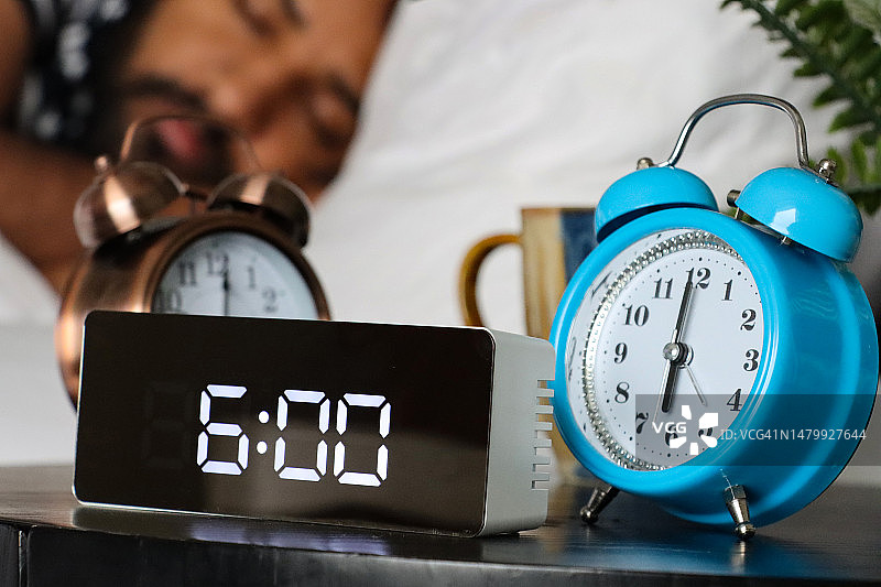 床头柜上三个闹钟特写，设置为早上6:00，清晨上班的睡眠男子，白色羽绒被和床单，数字和模拟时间，蓝色和金属钟图片素材