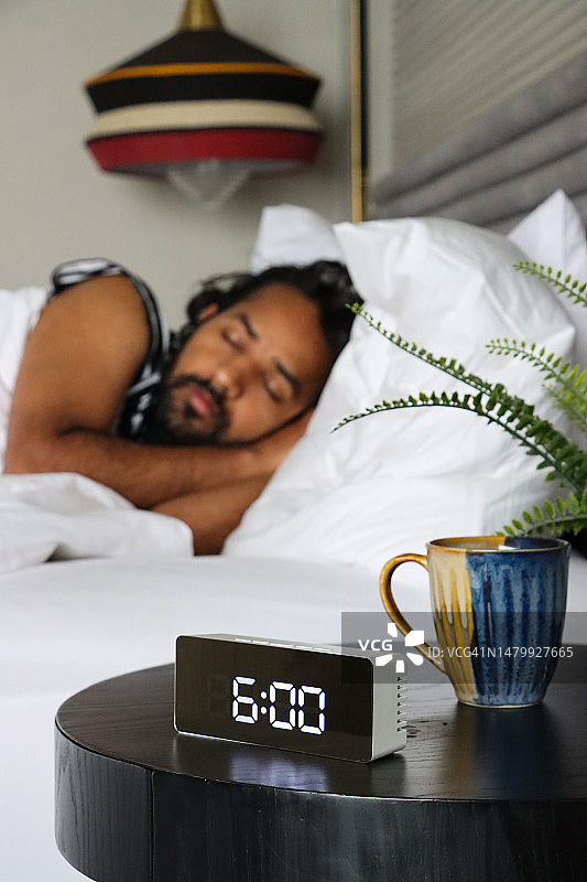 床头柜上的闹钟图片，设置为早上6:00，清晨上班的男人，白色羽绒被和床单，数字时间，桌子上的马克杯和假植物，竖版图片图片素材