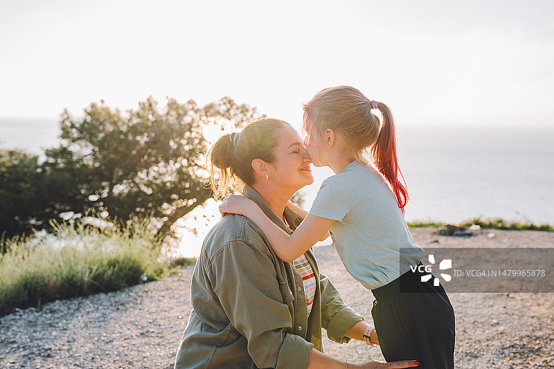母女探险：一起探索群山，阳光明媚的山景背景下，小女孩亲吻妈妈的鼻子图片素材