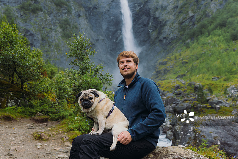 一名男子与狗在挪威风景秀丽的山谷瀑布前沉思的画像图片素材