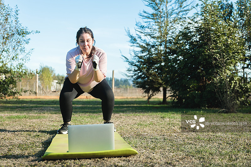 在瑜伽垫上做深蹲，观看电脑锻炼教程的年轻拉丁裔女子图片素材