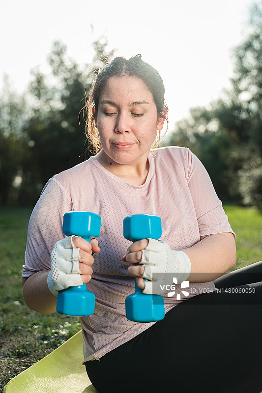 年轻超重女子在后院草地上用哑铃做腹部运动图片素材