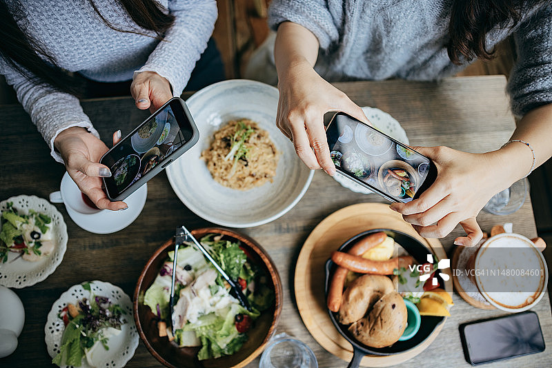 两个女孩在咖啡馆用餐，菜品上桌后用手机拍照并上传到社交媒体，分享快乐聚会图片素材