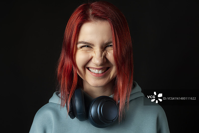 戴着蓝色超大连帽衫，用耳机享受音乐的迷人年轻红发女孩。 黑色背景。图片素材