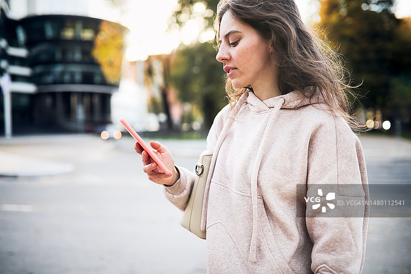 迷人的女子在小镇街头穿着运动帽衫，手持智能手机图片素材