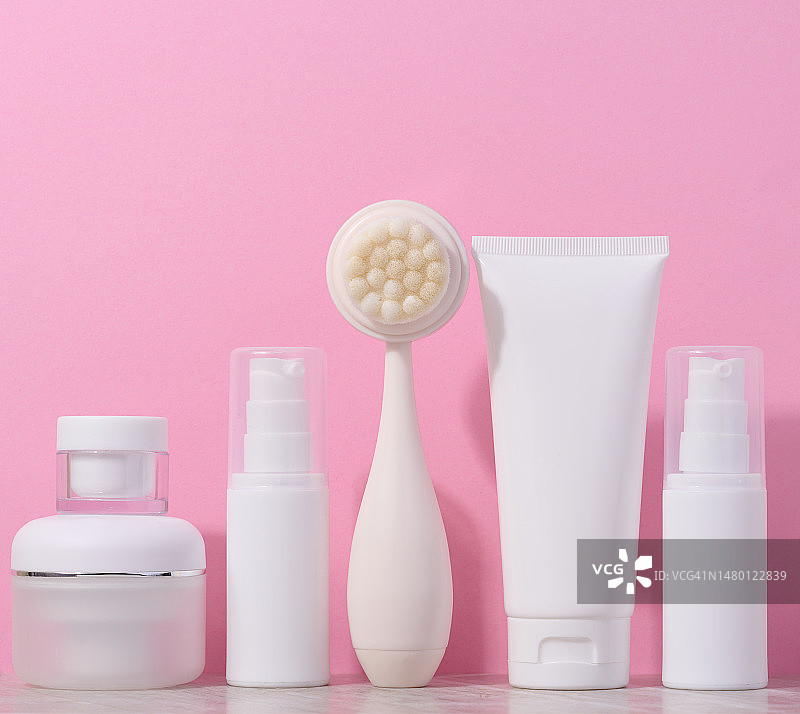 粉色背景上的白色塑料软管、面霜罐和面部清洁按摩刷图片素材