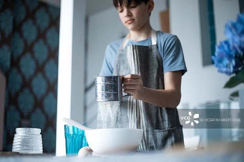 穿着厨房围裙的少年在厨房里筛面粉图片素材