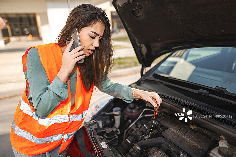 身穿反光衣服的年轻女子站在停在路上的抛锚汽车旁，打电话寻求帮助图片素材
