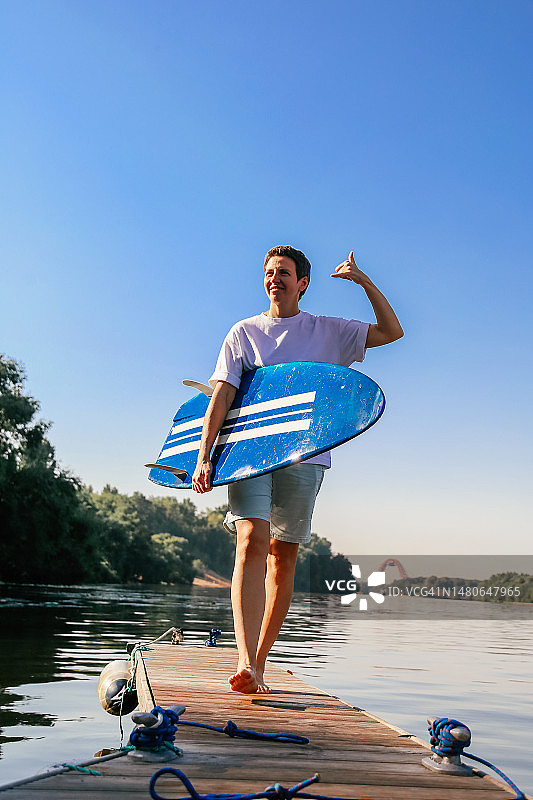 中年女性冲浪者走在木制码头上，拿着蓝色冲浪板，微笑并问候朋友图片素材