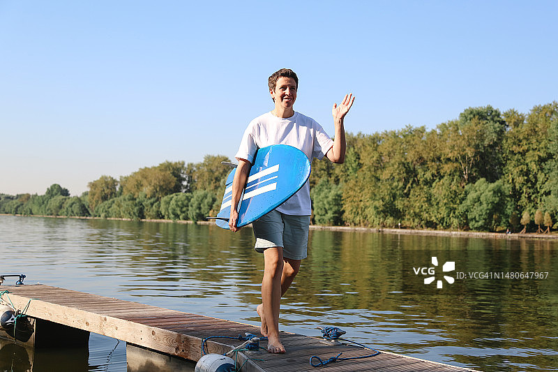 中年女性冲浪者手持蓝色冲浪板在木制码头上行走，并与朋友打招呼图片素材