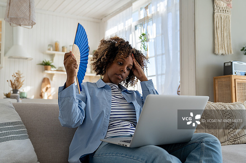 黑人女人在没有空调的情况下，坐在家里的沙发上扇扇子，中暑图片素材