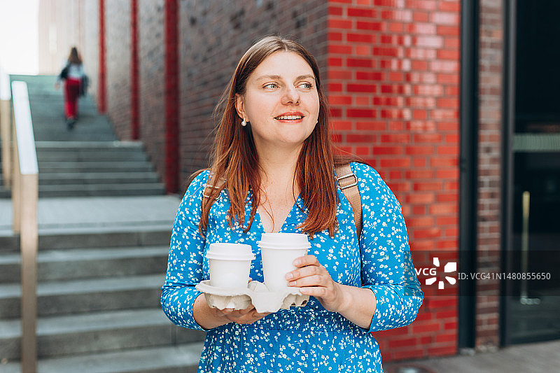 年轻女子身穿蓝色连衣裙在户外散步，快乐地拿着两杯外卖咖啡，城市背景，都市生活图片素材
