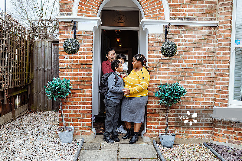 在英国的多种族亚裔和非洲裔家庭，站在前院的门廊上，男孩穿着校服去上学图片素材