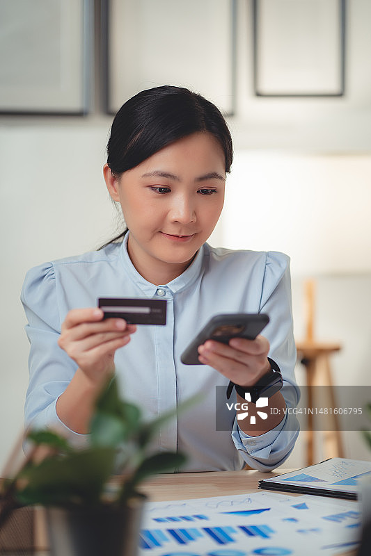 快乐的亚洲女人在家用智能手机进行网上支付，并持有信用卡进行网上购物图片素材