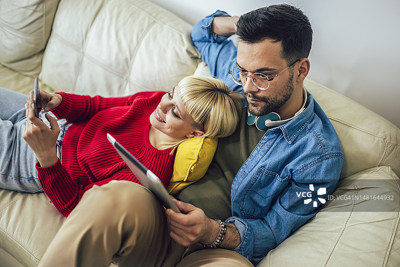 情侣在沙发上放松拥抱，使用平板电脑和手机图片素材