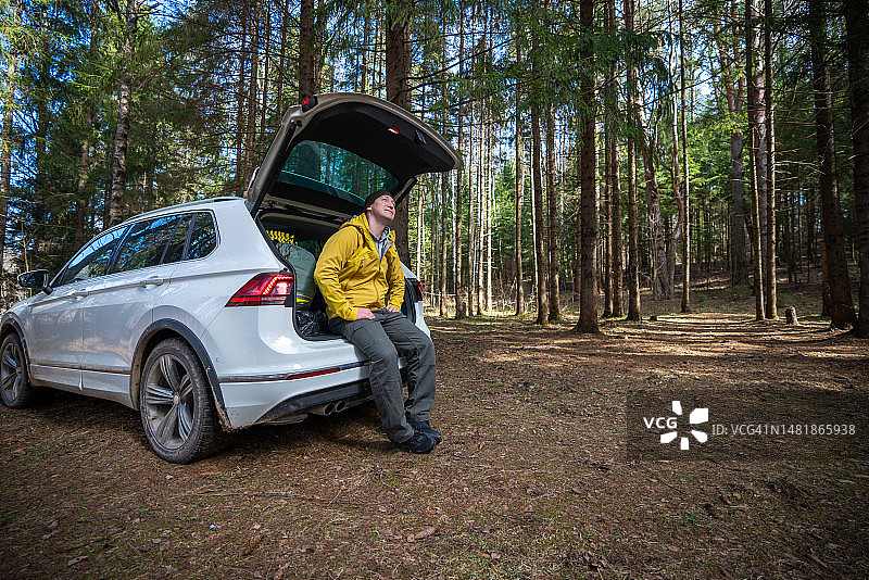 男人坐在汽车后备箱上，欣赏树林中露营地的景色图片素材