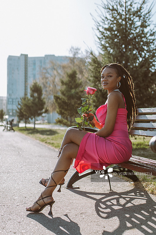 一位美丽的非裔美国年轻女性身穿粉色晚礼服坐在城市日落时的长椅上图片素材