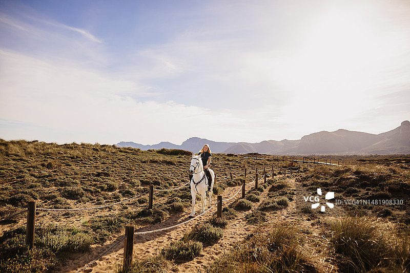 年轻的金发女子在靠近海边的沙丘上骑着她的白马图片素材
