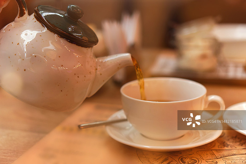 在舒适的茶馆里，手将红茶倒入白色陶瓷杯中，特写镜头呈现天然热饮，正面视角，高角度拍摄图片素材