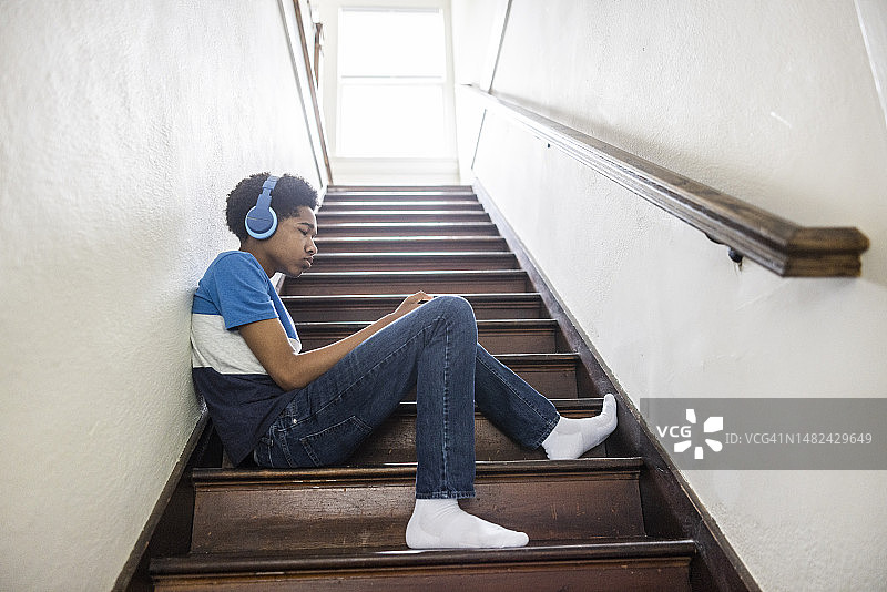 青少年男孩在家中楼梯上用无线耳机和智能手机图片素材