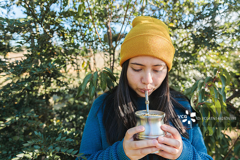 年轻女人在公园里喝马黛茶，背景是茂盛的树叶图片素材