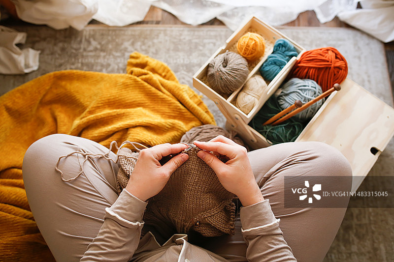女人在舒适的家中用一盒纱线和木制针织针编织棕色物品。顶视图，无法辨认的人。图片素材