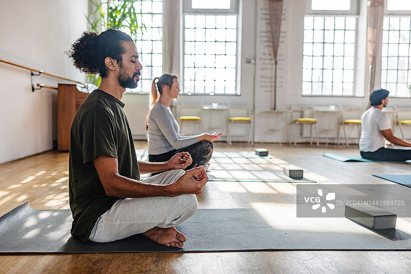 瑜伽课参与者坐在瑜伽垫上，在带大窗户的漂亮工作室里冥想图片素材