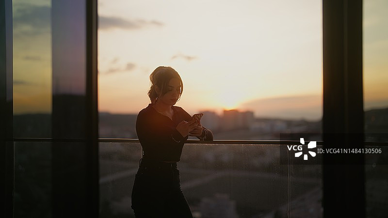 日落时分在阳台上使用智能手机的年轻Z世代女性图片素材