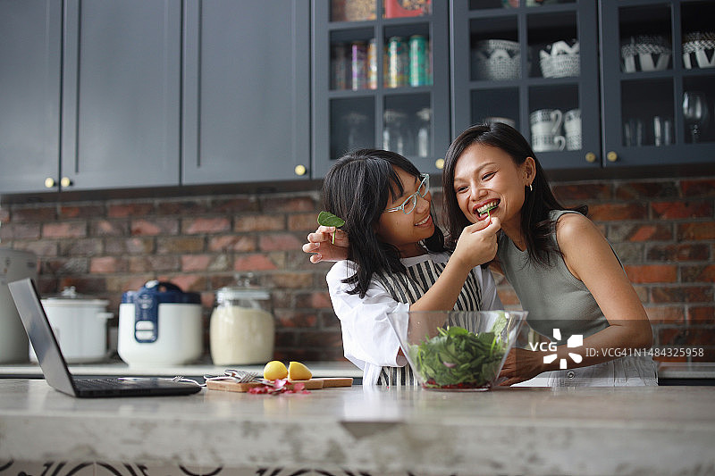 一位快乐的亚洲母亲和女儿在厨房里使用笔记本电脑，一边拌沙拉一边玩耍图片素材