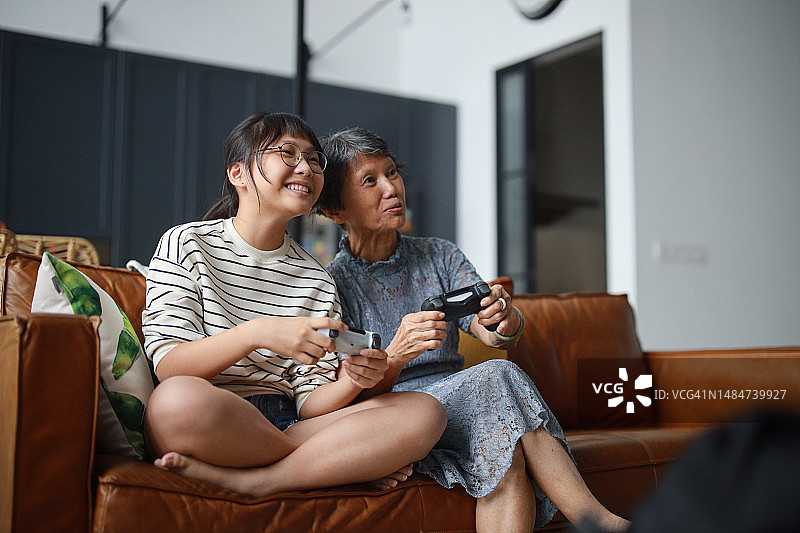 快乐的亚洲祖母和孙女坐在客厅的沙发上，在家一起玩电子游戏，温馨的场景体现了快乐的退休理念图片素材
