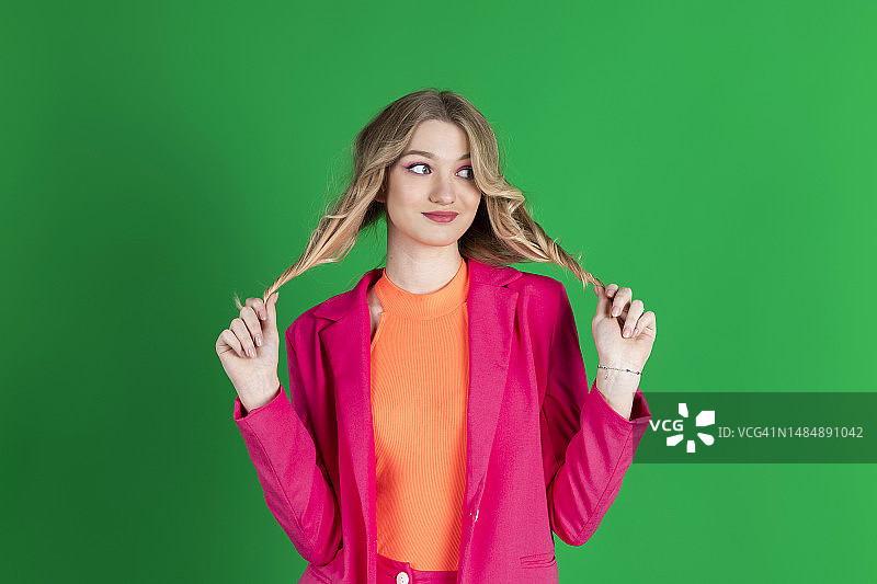 穿着粉色西装，看向远方，拉着头发的年轻卷发金发女子，绿色背景图片素材